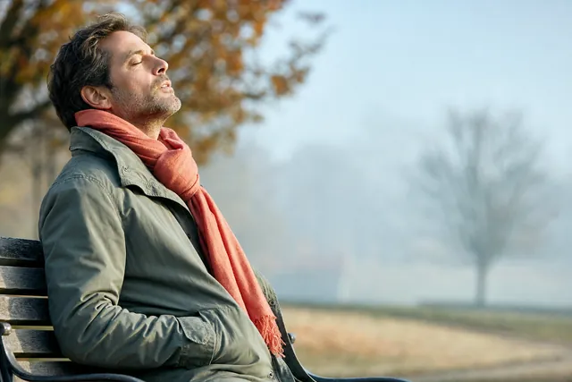A man on a park bench on an autumn morning tilts his head back with eyes closed, letting out a long slow breath